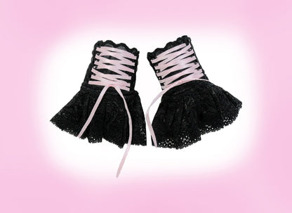 Sweet Lolita Manschetten aus Spitze in schwarz mit Zier-Schnürung & Röckchen für Cyber, Punk, Harajuku, Gothic Outfits. Handgefertigt in Deutschland. Hohe Qualität. Super Preis-Leistungs-Verhältnis. Keine billige China Ware. Die kleinen Schätze sind flatlay flach liegend nebeneinander fotografiert auf einem Rosa Hintergrund mit weißem Schatten. Die Schnürung schnürt sich mit einem schmalen Satinband wie ein Bondage vom Manschetten-Teil zum Röckchen hinunter.