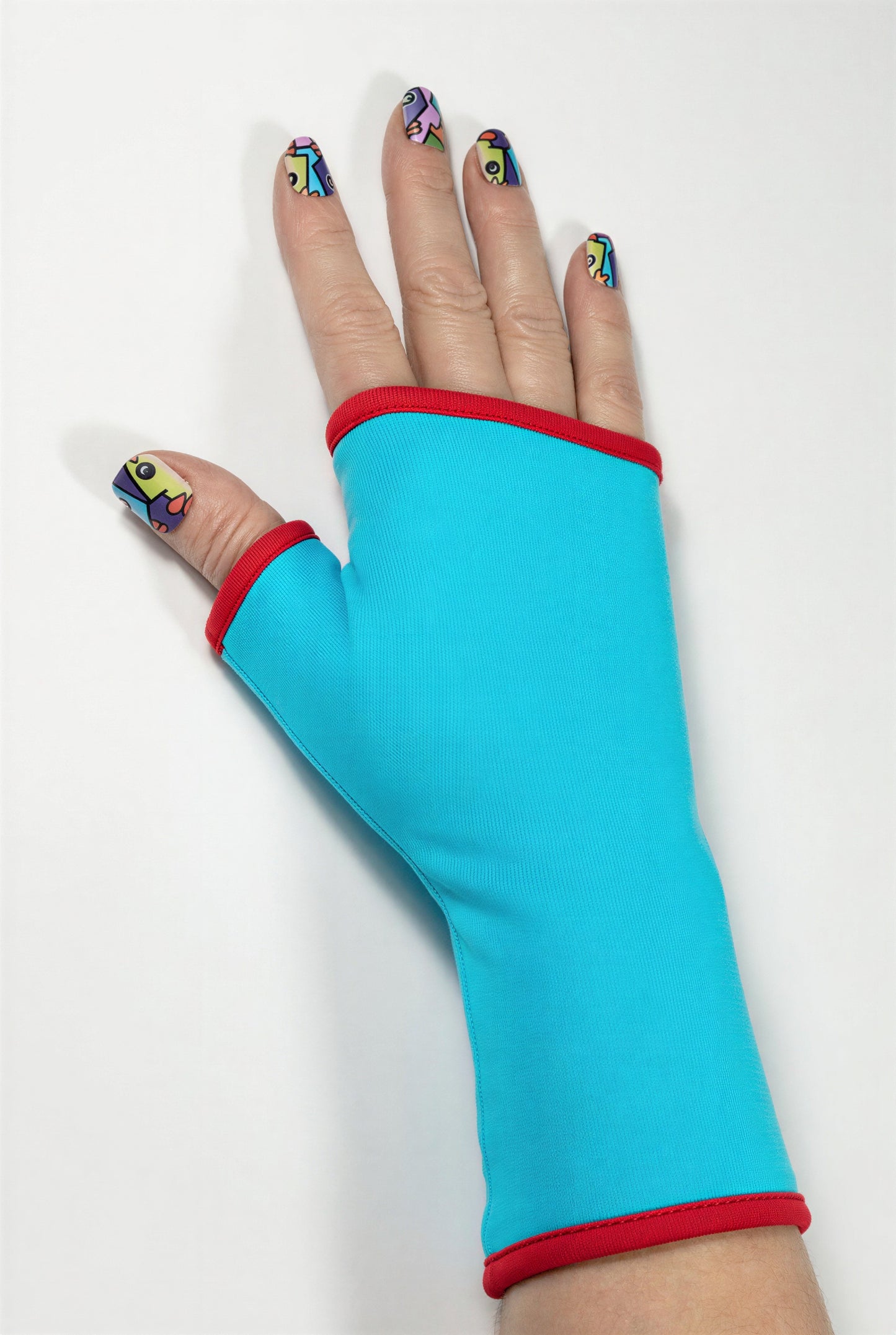Einzel Basic Handschuh ohne Finger, fotografiert an einer Damenhand, die auf dem Tisch liegt. Bunt lackierte Fingernägel, sehr stylisch und exklusiv. Aus Baumwolle Jersey mit Paspel, gefüttert. Oberstoff türkis, Futter rot.