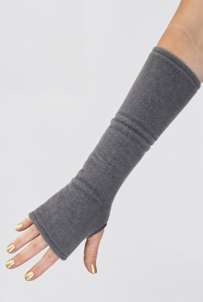 eine Fleece Armstulpe dargestellt an einem Frauenarm. Die Stulpen reichen bis an die Fingerknöchel über die Hand, ohne Finger, fingerlos, aber mit Daumenloch zum rein schlüpfen und bis über den Unterarm hinauf fast bis zur Elle. Material ist flauschig, fluffig und wärmend. Farbe grau.